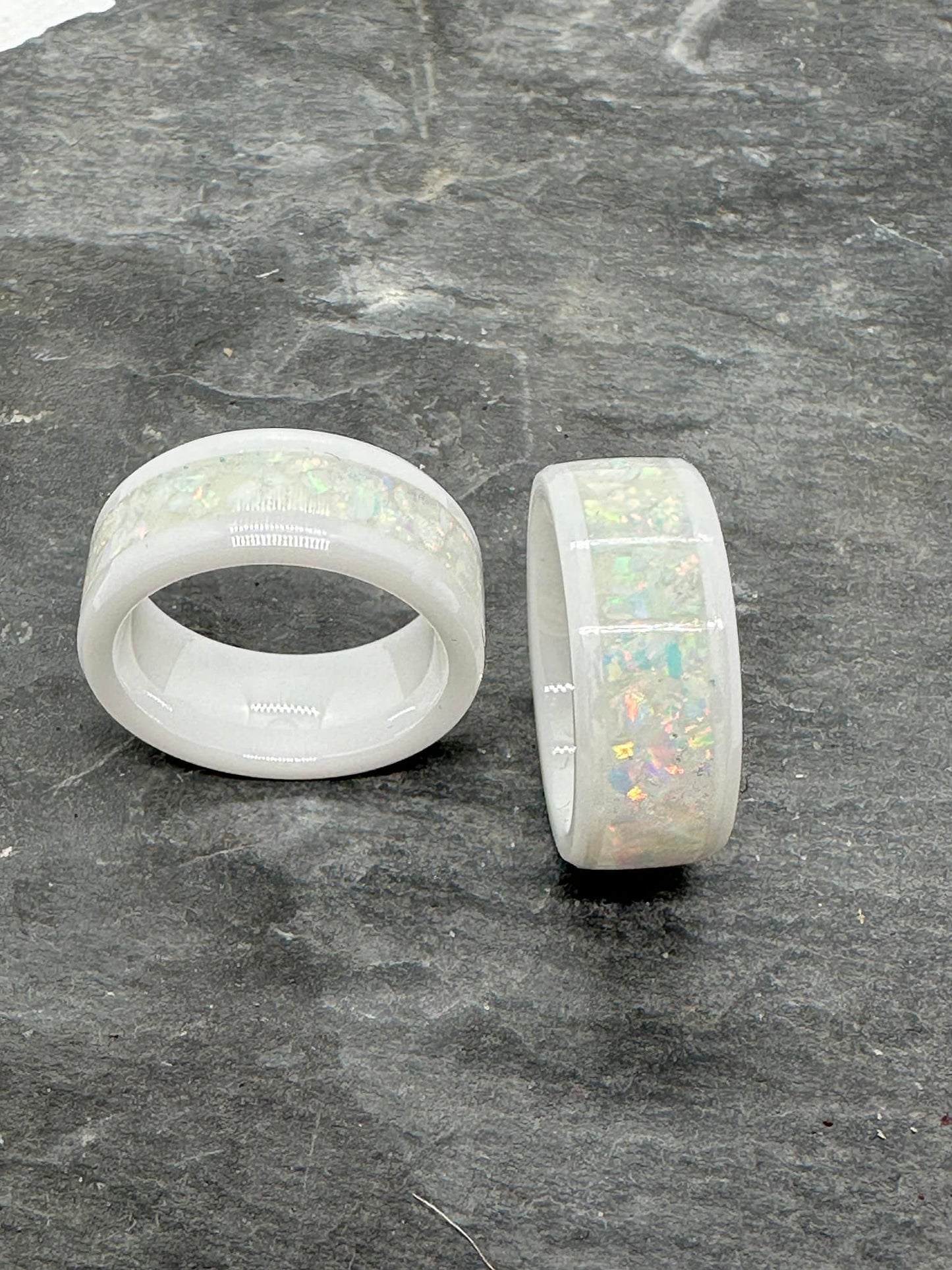 Zwei White Lady Ringe aus weißer Keramik mit eingelegten weißen Opalen. Das irisierende Farbspiel der Opale schimmert sanft im Kontrast zur matten Keramik. Alt-Text (EN): Two White Lady rings made of white ceramic with inlaid white opals. The iridescent play of colors of the opals shimmers gently against the matte ceramic.
