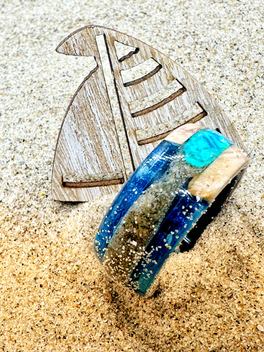 Handgefertigter Ring, gefüllt mit Sand und kleinen Elementen vom Strand. Transparente Harzschicht bewahrt die Struktur des Strandes und macht ihn als tragbare Erinnerung erlebbar.

Handmade ring filled with sand and small elements from the beach. A transparent resin layer preserves the texture of the shore, turning it into a wearable memory.