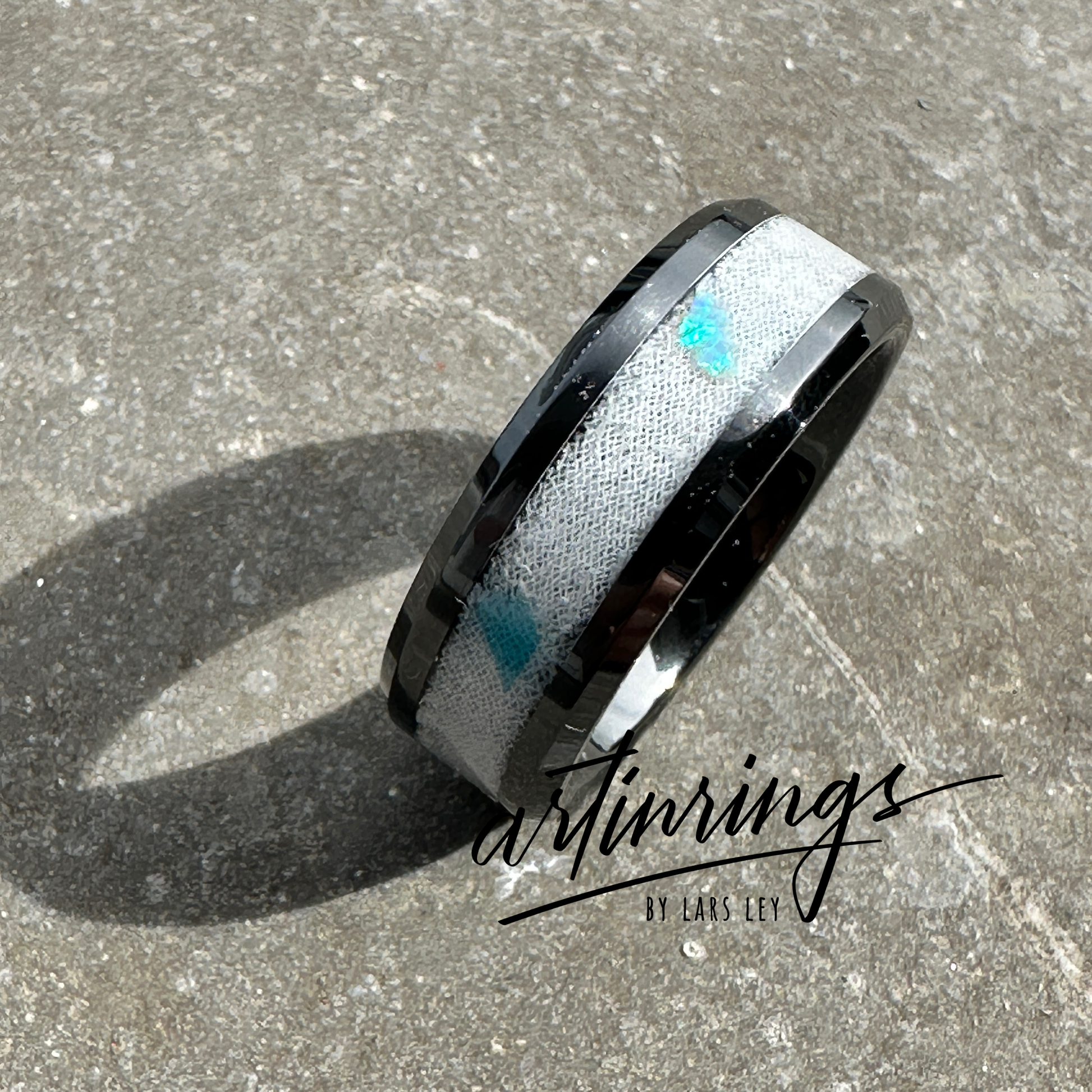 Schwarzer Keramikring, gefüllt mit weißem Brautkleidstoff und blauen Opalen. Der helle Stoff im Kontrast zum tiefen Schwarz der Keramik wird von den schimmernden Blautönen der Opale akzentuiert – ein Ring voller Symbolik von Liebe und Ewigkeit.

Black ceramic ring filled with white bridal dress fabric and blue opals. The light fabric contrasts with the deep black ceramic, highlighted by the shimmering blue tones of the opals – a ring full of symbolism of love and eternity.