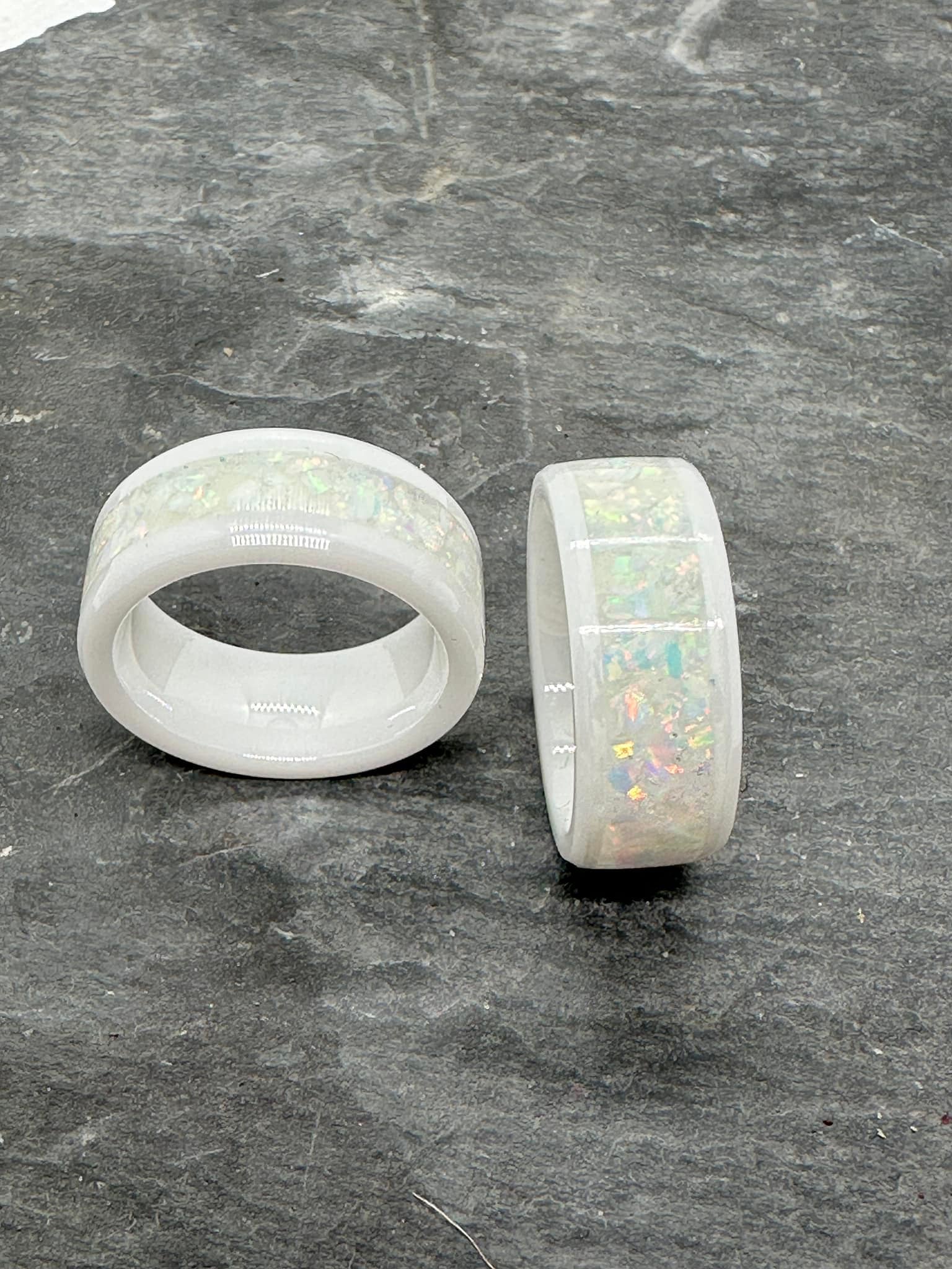 Zwei White Lady Ringe aus weißer Keramik mit eingelegten weißen Opalen. Das irisierende Farbspiel der Opale schimmert sanft im Kontrast zur matten Keramik.  Alt-Text (EN): Two White Lady rings made of white ceramic with inlaid white opals. The iridescent play of colors of the opals shimmers gently against the matte ceramic.
