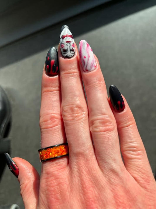 Hand mit langen Halloween-Nägeln in Schwarz, Rot und Weiß sowie Horror-Motiv. Getragen wird der Ring Jack O’ aus schwarzer Keramik mit intensiv orangefarbenen Opalen, die im Licht glühen.  Alt-Text (EN): Hand with long Halloween-themed nails in black, red, and white with a horror design. Worn is the Jack O’ ring made of black ceramic with intense orange opals that glow in the light.