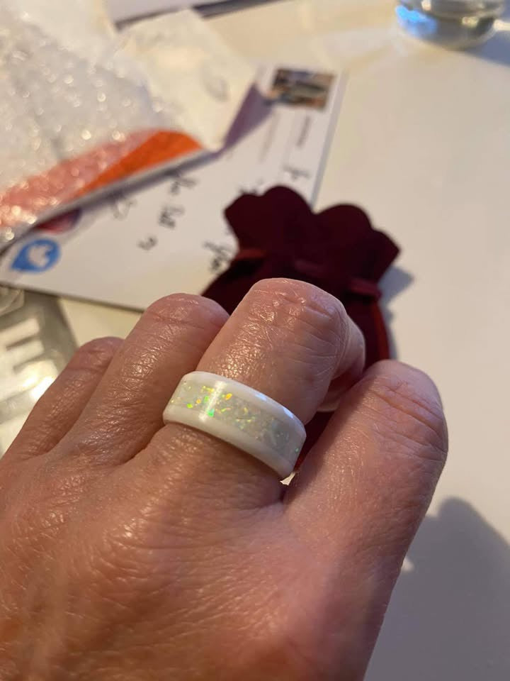 Hand trägt den Ring White Lady aus weißer Keramik mit eingelegten weißen Opalen. Die Opale funkeln dezent in Gelb- und Grüntönen und heben sich elegant vom hellen Keramikband ab.  Alt-Text (EN): Hand wearing the White Lady ring made of white ceramic with inlaid white opals. The opals sparkle subtly in yellow and green tones, standing out elegantly against the bright ceramic band.