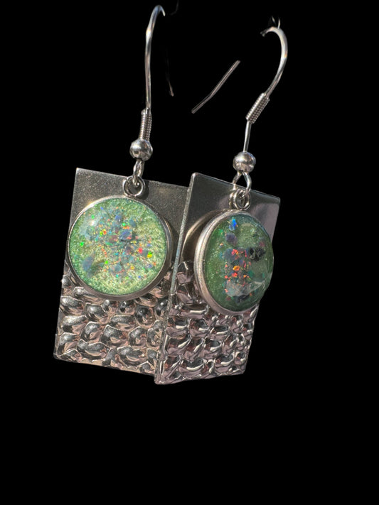 Handgefertigte Dangle-Ohrringe mit rechteckigem Silberrahmen und strukturierter Oberfläche. Eingesetzt sind runde Elemente mit schimmernden grünen Opalen, die in verschiedenen Farben funkeln.

Handmade dangle earrings with a rectangular silver frame and textured surface. Round elements with shimmering green opals are set in, sparkling with multicolor reflections.