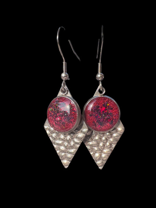 Handgefertigte Dangle-Ohrringe mit rautenförmigen Silberrahmen und strukturierter Oberfläche. Eingesetzt sind runde Elemente mit schimmernden roten Opalen, die in verschiedenen Farben funkeln.

Handmade dangle earrings with a square silver frame and textured surface. Round elements with shimmering red opals are set in, sparkling with multicolor reflections.